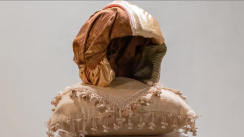 A brown patterned and light gold velvet cloth cover a helmet like shape, it sits upon a soft beige pillow with many tassels hanging down.