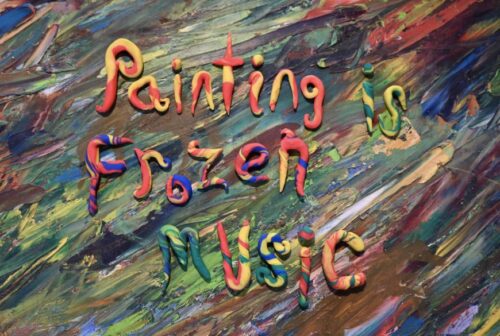 On a multicolored background the words in sculpey clay say Painting is Frozen Music. Painting is in red and yellow. Is in green and yellow. Frozen in red blue and yellow. and music in all the colors changing in each letter. 